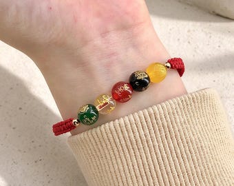 Puede incluir: Una pulsera con cinco cuentas redondas multicolores en verde, transparente, rojo, negro y amarillo, cada una decorada con escritura dorada. Las cuentas están ensartadas en un cordón trenzado rojo con pequeños detalles metálicos dorados.