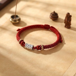 Puede incluir: Una pulsera de cordón trenzado rojo con una cuenta cilíndrica central de tono plateado con textura tejida, flanqueada por dos cuentas esféricas de color rojo oscuro y dos pequeños acentos dorados, con cierre de nudo corredizo.