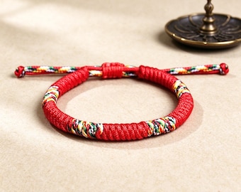 Puede incluir: Una pulsera de cordón trenzado rojo con secciones de hilo tejido multicolor en amarillo, verde y blanco. La pulsera tiene un cierre de nudo corredizo ajustable con dos extremos que también presentan el mismo patrón multicolor.