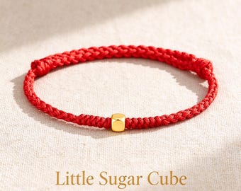 Puede incluir: Una pulsera de cordón trenzado rojo con una cuenta cúbica de tono dorado pulido en el centro. El texto debajo dice: Little Sugar Cube ... Sweet or Bold, Lightly Tempting ... Este accesorio tiene un diseño sencillo con cierre de nudo.