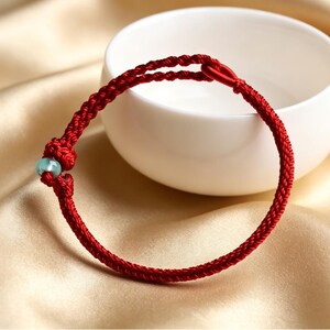 May include: A red braided cord bracelet featuring a single light green bead and a knotted closure. The bracelet is constructed from a thin, woven red string that forms a circular band, with a small, smooth, pale green bead secured by a knot.