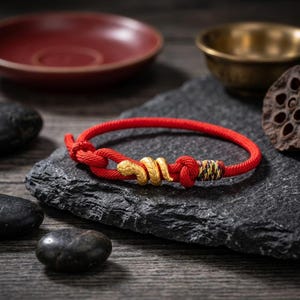 May include: A red rope bracelet with gold-colored snake and ring accents. The bracelet is knotted and rests on a dark gray stone surface. Other objects in the background include a red bowl and a small brass bowl.