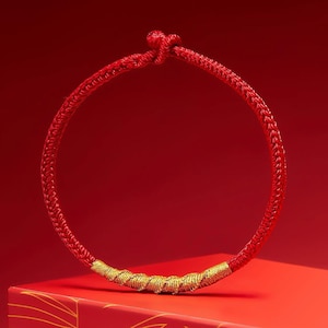 May include: A red braided bracelet with a gold-colored accent. The bracelet is circular, with a knot at the top. The gold accent is a series of tightly wound strands. The background is a solid red.