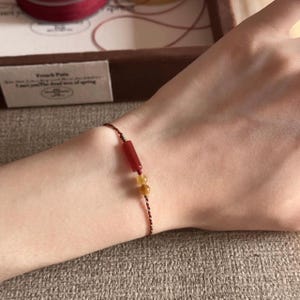 以下が含まれることがあります： 細いダークレッドのツイストコードを使用した繊細なブレスレット。円筒形の赤いビーズ1つと、半透明の黄色い球状ビーズ2つがあしらわれており、コードには小さな金属製のアクセントビーズが固定されています。