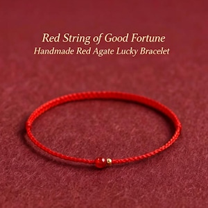 Puede incluir: Una pulsera de cordón rojo fino con una pequeña cuenta de ágata roja y una diminuta cuenta de metal dorado. El texto sobre la pulsera dice: Red String of Good Fortune, Handmade Red Agate Lucky Bracelet. El diseño es sencillo y circular.