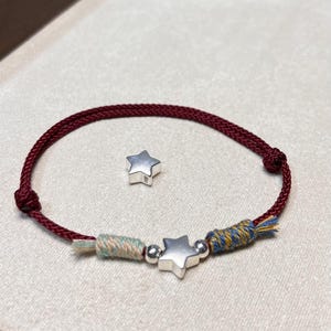 Peut inclure: Un bracelet en cordon rouge foncé avec une breloque étoile argentée centrale, entourée de deux petites perles argentées et de deux sections de fils multicolores enroulés. Une breloque étoile argentée isolée est placée au-dessus.