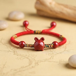 Op de afbeelding: Een rood koordarmband met een centrale rode keramische bedel in de vorm van een muis met een klein reliëfkarakter aan de voorzijde. De armband bevat twee rode bolvormige kralen en secties van gevlochten geel, groen en zwart garen.