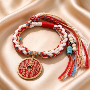 Op de afbeelding: Een gevlochten armband met witte, rode en blauwe koorden en een ronde metalen hanger. De hanger heeft een rood middenstuk met gouden Chinese karakters en een gouden rand. De armband bevat houten kralen, turkoois kralen en lange kwastjes.