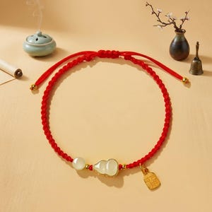 May include: A red braided cord bracelet featuring a central white jade-like gourd charm, a smooth round white bead, and a small rectangular gold-toned metal pendant with engraved characters, accented by tiny gold-toned beads along the cord.