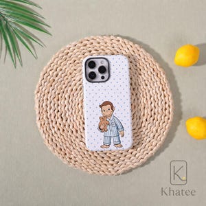 May include: A white smartphone case featuring a pattern of small blue dots and a central illustration of a brown monkey wearing blue and white striped pyjamas while holding a small brown teddy bear. The case has a black camera lens cutout.