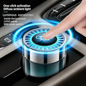 May include: A car interior accessory with the Audi logo, featuring a glowing blue light. The device has a silver and black finish and is being activated by a finger. Text on the image reads: One-click activation, Diffuse ambient light, Luminous oanelinteri, Inside the luminous LOGO panel, With press switch.