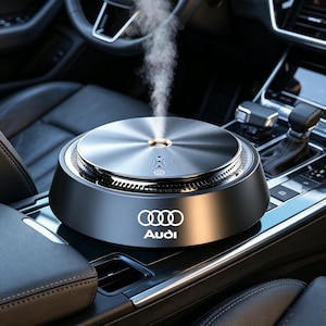 Puede incluir: Un ambientador para coche negro y plateado con el logotipo de Audi. El dispositivo emite un chorro de vapor. El interior del coche es visible en el fondo, incluyendo el volante, la palanca de cambios y los asientos.