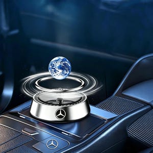 Puede incluir: Un ambientador de coche plateado con un diseño de globo terráqueo. El ambientador tiene un diseño circular con un logotipo de Mercedes-Benz. El globo es azul y blanco, y el ambientador está colocado en el salpicadero de un coche.