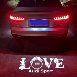 Op de afbeelding: Zilveren auto met rode achterlichten. De kentekenplaat van de auto is blauw. Onder de auto staat het woord "LOVE" in het wit, met het Audi-logo en de woorden "Audi Sport".