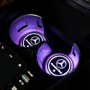 May include: A close-up of two car cup holders with a purple glow. Each cup holder has a circular insert with the Mercedes-Benz logo in white. The car's control panel is visible in the lower left corner.