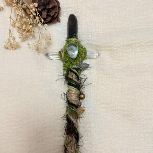 May include: A decorative wand featuring a dark smoky quartz point at the top, wrapped in natural jute twine and green moss. It includes a clear crystal accent, a small silver-toned leaf charm, and a tiny brass bell, with horizontal clear crystals.