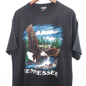 Op de afbeelding: Zwart T-shirt met een afbeelding van een Amerikaanse zeearend die over een waterval en bos vliegt. Het woord "TENNESSEE" is in het wit gedrukt. Het shirt heeft een ronde hals en korte mouwen. Het merk "SIGNAL" is zichtbaar.