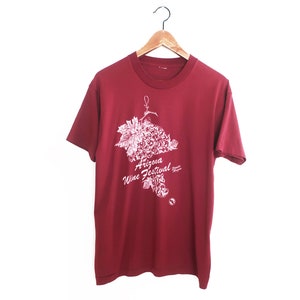 Op de afbeelding: Een rood T-shirt met een witte afbeelding van een tros druiven en de tekst "Arizona Wine Festival".