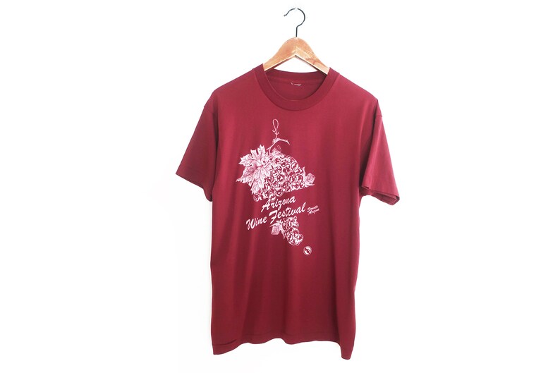 Op de afbeelding: Een rood T-shirt met een witte afbeelding van een tros druiven en de tekst "Arizona Wine Festival".
