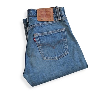 May include: Blue denim jeans with a classic five-pocket design. The jeans feature a red Levi's tag on the back pocket and a brown leather patch with the Levi's logo. The jeans are folded and displayed on a white background.