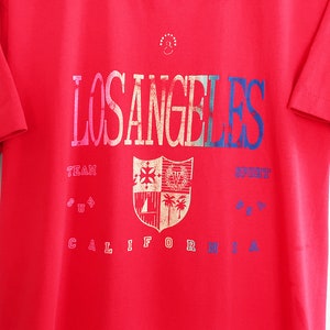 May include: Red t-shirt with a vintage Los Angeles graphic featuring a shield with a crest and palm trees. The text reads "Los Angeles Team Sport California".