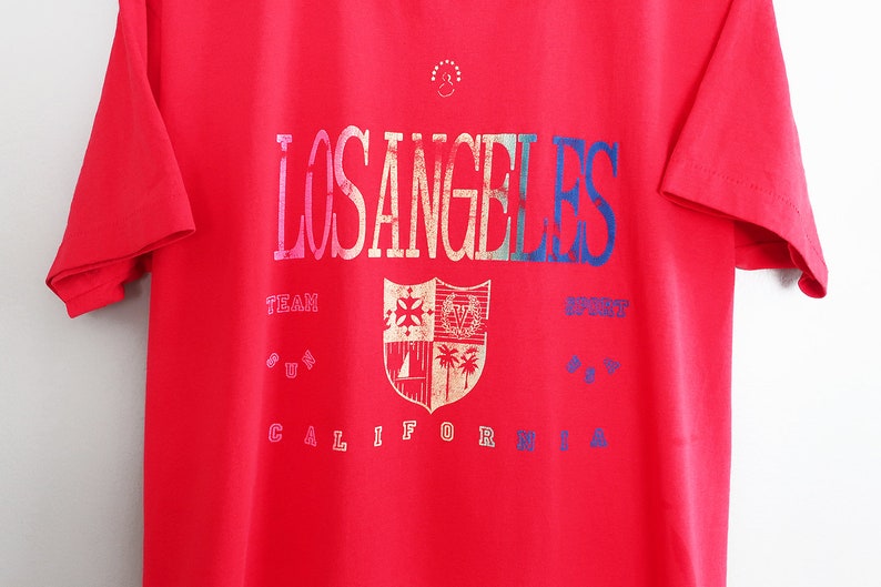 May include: Red t-shirt with a vintage Los Angeles graphic featuring a shield with a crest and palm trees. The text reads "Los Angeles Team Sport California".
