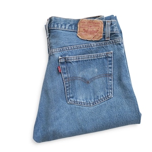 May include: A pair of folded blue denim jeans featuring a tan leather patch on the waistband with the text Levi Strauss & Co. and a small red tab on the back pocket. The jeans have a classic five-pocket design with visible stitching and metal rivets.