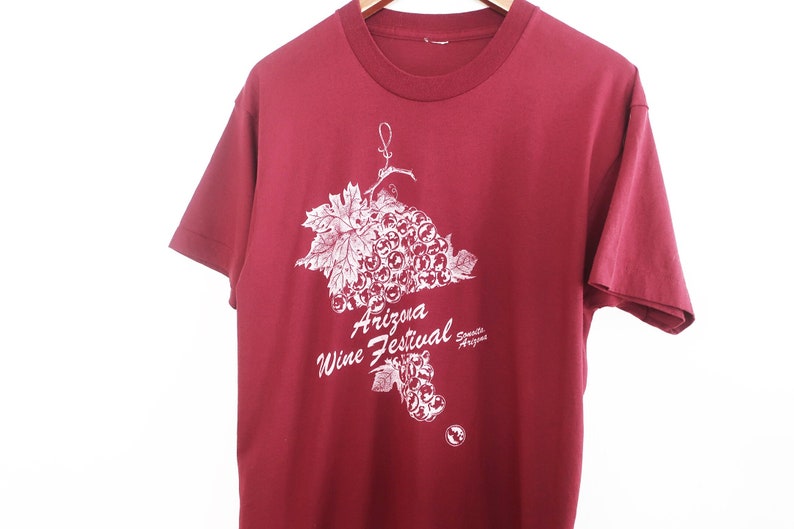 Op de afbeelding: Een rood T-shirt met een witte afbeelding van een tros druiven en de tekst "Arizona Wine Festival, Scottsdale Arizona".