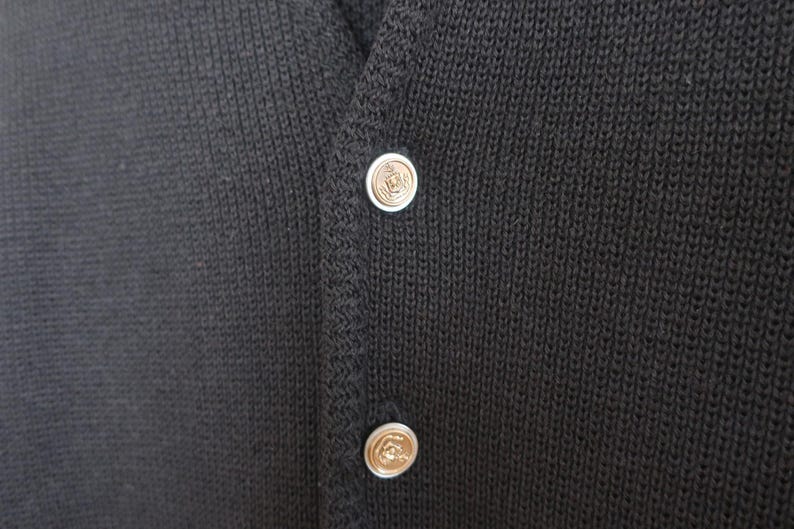 May include: Close-up of a dark gray knitted cardigan with two decorative buttons. The buttons are silver with a gold-colored center. The cardigan has a V-neck and a textured knit pattern.