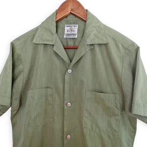 Puede incluir: Camisa verde oliva de manga corta con dos bolsillos delanteros y cuello. La camisa está abotonada y colgada de una percha de madera. La etiqueta dice "Permanent Press" y "John Rulla".