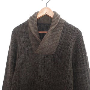 Könnte beinhalten: Ein brauner Strickpullover mit Schalkragen, der an einem Holzbügel hängt. Der Pullover hat eine strukturierte Optik und einen dunkleren braunen Kragen. Der Hintergrund ist schlicht weiß.