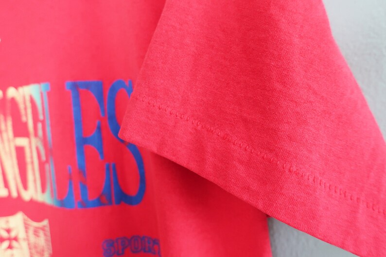 May include: Red t-shirt with a blue and white "Los Angeles" graphic print. The shirt has a ribbed crew neck and short sleeves.