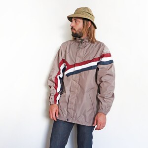 Pu&ograve; includere: Una giacca beige con chiusura a zip e un design a strisce bianche, rosse e blu. La giacca ha un cappuccio e un cordoncino in vita.