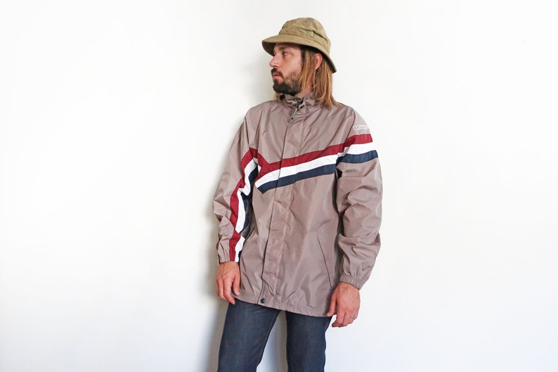 Pu&ograve; includere: Una giacca beige con chiusura a zip e un design a strisce bianche, rosse e blu. La giacca ha un cappuccio e un cordoncino in vita.