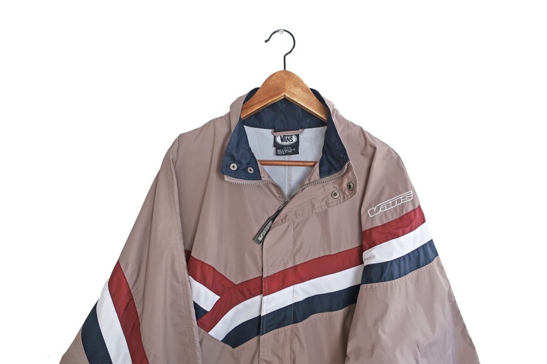 Pu&ograve; includere: Una giacca a righe beige, blu, rossa e bianca con chiusura a zip e un logo Vans sulla manica destra.
