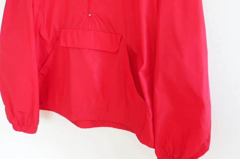 May include: A red windbreaker jacket with a zip closure and a kangaroo pocket. The jacket has long sleeves and a drawstring hood.