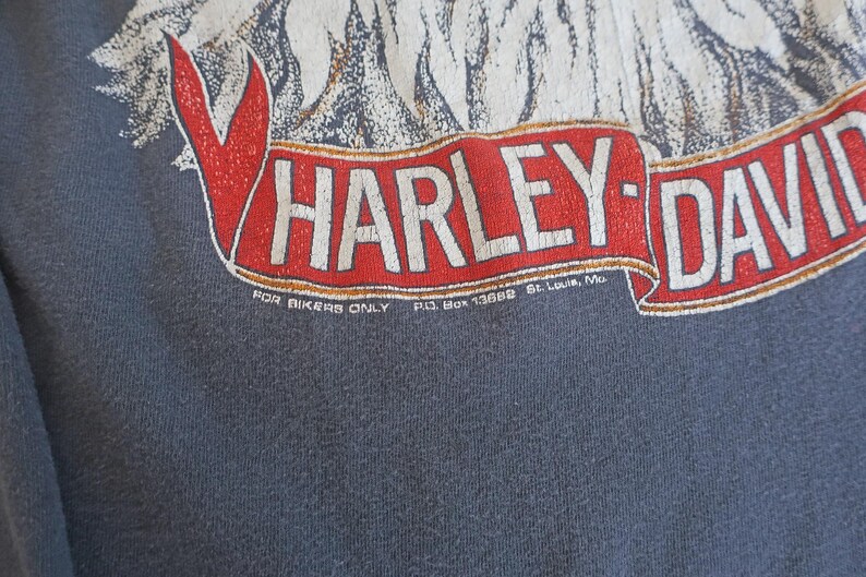 May include: A dark blue t-shirt with a white eagle graphic and a red and white Harley-Davidson logo. The text "FOR BIKERS ONLY P.O. BOX 13888 St. Louis, Mo." is printed below the logo.