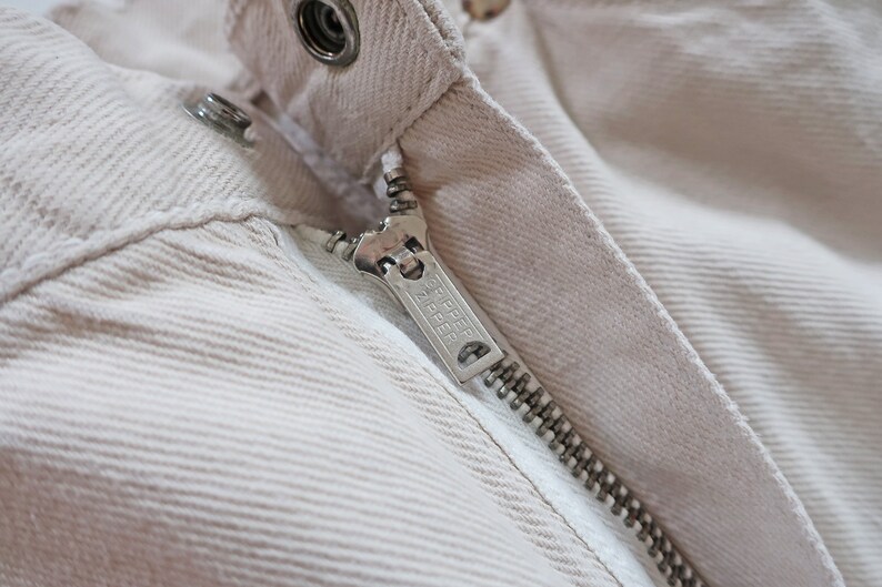 K&ouml;nnte beinhalten: Nahaufnahme eines Rei&szlig;verschlusses an einer beige Cordjacke. Der Rei&szlig;verschluss-Schieber tr&auml;gt den Schriftzug "YKK".