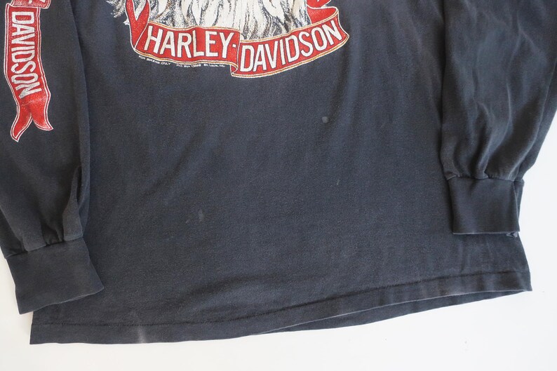 May include: Black long-sleeve t-shirt with a Harley-Davidson eagle graphic and red ribbon accents. The shirt has a distressed look and is made of a soft, comfortable fabric.