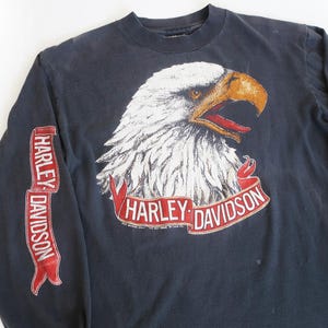 May include: A black long-sleeve t-shirt with a white eagle graphic and red Harley-Davidson banner. The eagle has a red beak and yellow eyes.