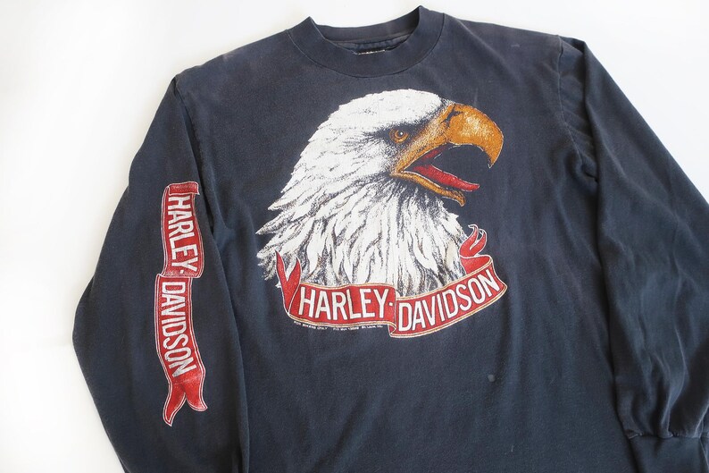 May include: A black long-sleeve t-shirt with a white eagle graphic and red Harley-Davidson banner. The eagle has a red beak and yellow eyes.