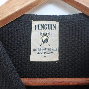 May include: Close-up of a black knitted sweater with a label that reads "PENGUIN" above a crest design. The label also states "SEATTLE KNITTING MILLS ALL WOOL M". The sweater is hanging on a wooden hanger.