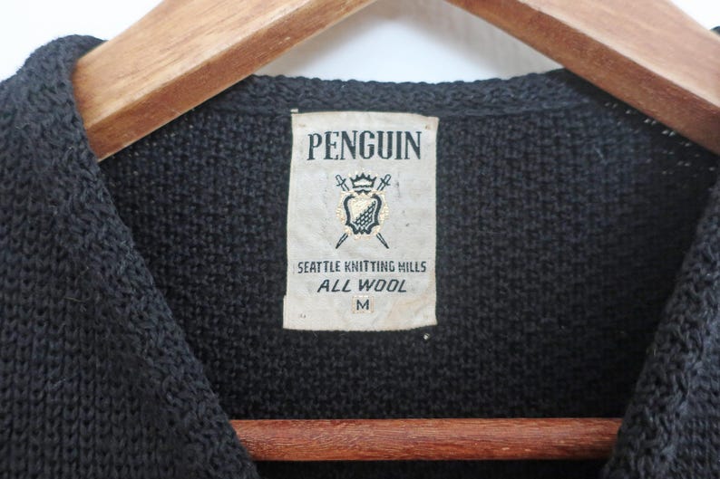 May include: Close-up of a black knitted sweater with a label that reads "PENGUIN" above a crest design. The label also states "SEATTLE KNITTING MILLS ALL WOOL M". The sweater is hanging on a wooden hanger.