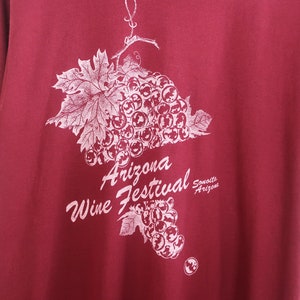 Op de afbeelding: Een maronkleurig T-shirt met een witte afbeelding van een tros druiven en de tekst "Arizona Wine Festival Sonoita Arizona".