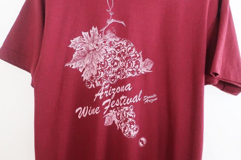 Op de afbeelding: Een maronkleurig T-shirt met een witte afbeelding van een tros druiven en de tekst "Arizona Wine Festival Sonoita Arizona".
