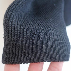 May include: Close-up of a black knit fabric sleeve cuff. The cuff is ribbed and shows a small hole. The fabric appears to be a dark, solid color, possibly wool or a similar material. The background is a plain white.