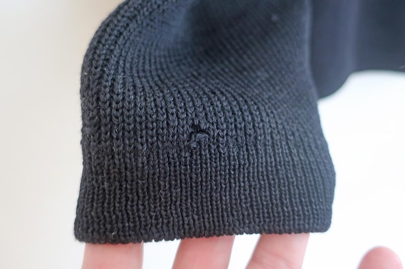 May include: Close-up of a black knit fabric sleeve cuff. The cuff is ribbed and shows a small hole. The fabric appears to be a dark, solid color, possibly wool or a similar material. The background is a plain white.