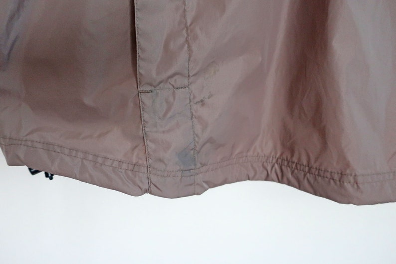 Pu&ograve; includere: Una giacca di nylon marrone con chiusura a zip e una piccola tasca sul lato sinistro. La giacca ha un cordoncino in fondo e una cucitura al centro della schiena.