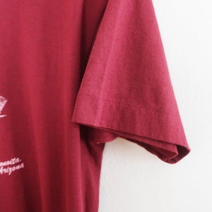 Op de afbeelding: Een maroon t-shirt met een wit druivenrankenontwerp en de tekst "Arizona Wine Festival" gedrukt op de voorkant.