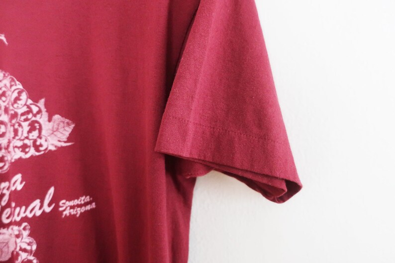 Op de afbeelding: Een maroon t-shirt met een wit druivenrankenontwerp en de tekst "Arizona Wine Festival" gedrukt op de voorkant.